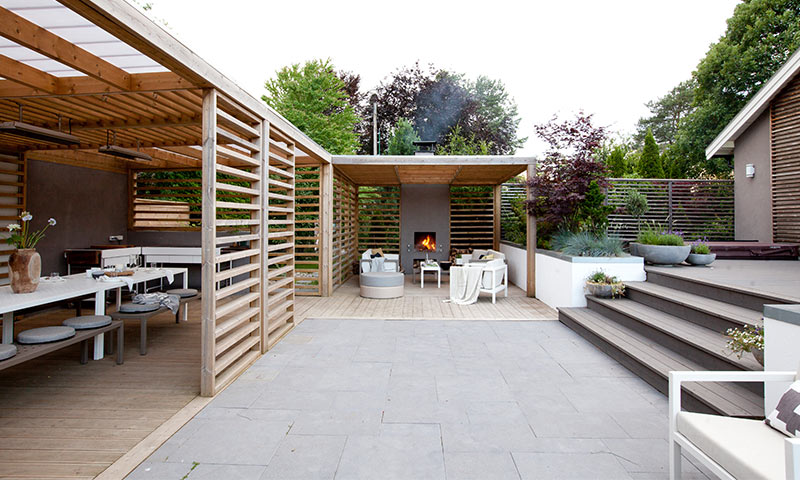 Moderne uteplass med lysegrå steinflater, trepergola, peis og hvite sittemøbler.