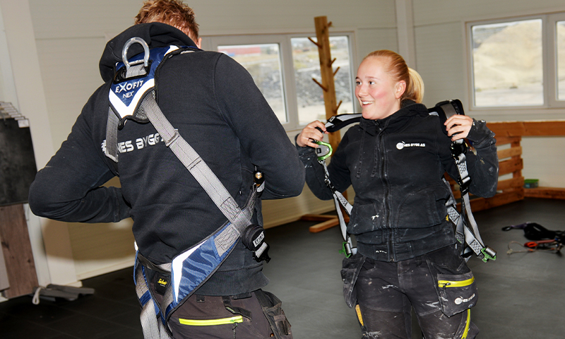 To personer i svarte arbeidsklær fester blågrå fallsikringsseler i lyst verkstedrom.