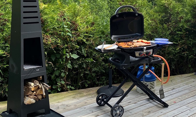 Sort gassgrill på treterrasse med kjøtt og pølser, blå tallerkener, vedstabel og grønn hekk