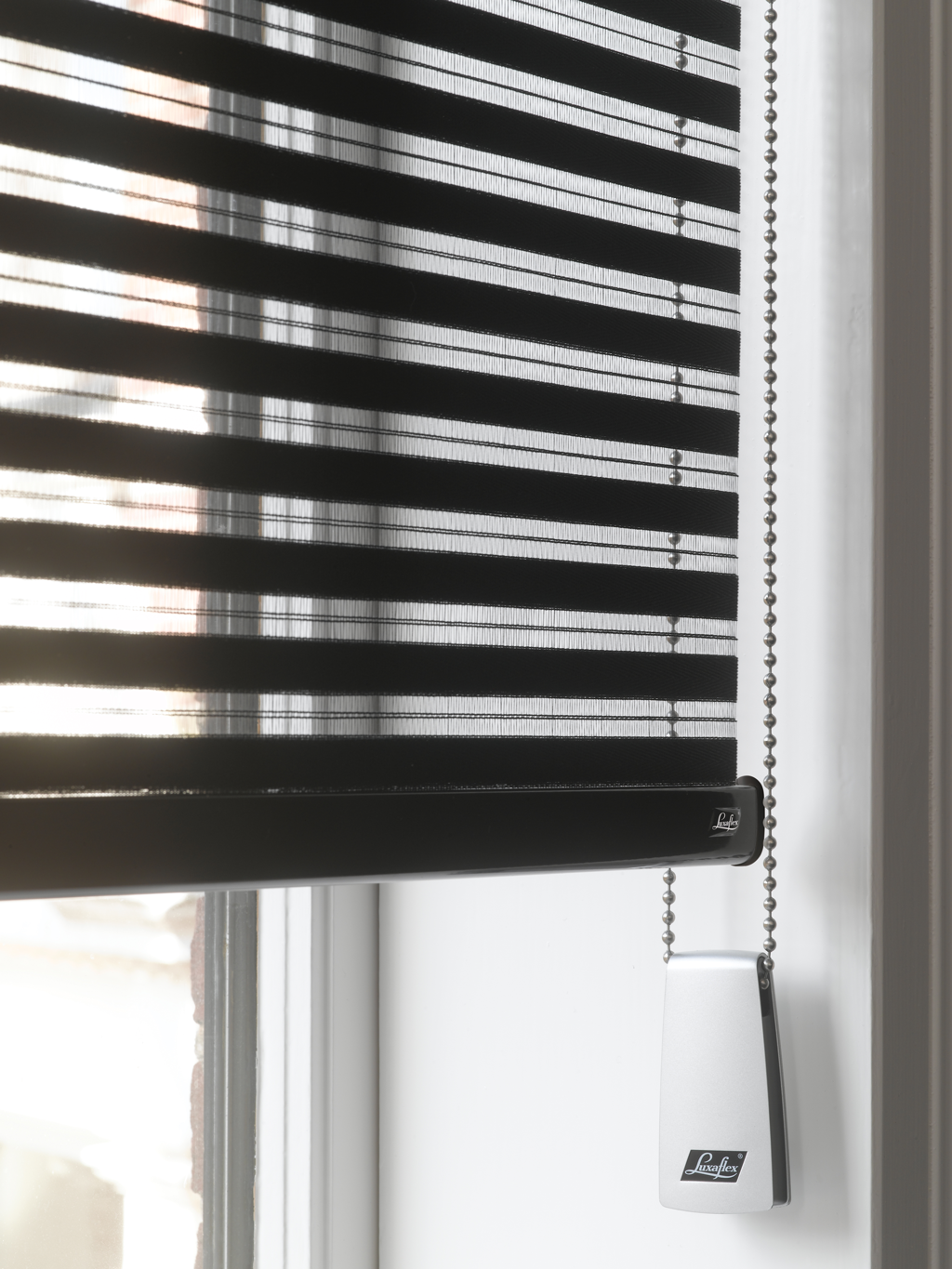 Svart Luxaflex persienne med horisontale lameller og kulekjede foran lyst vindu