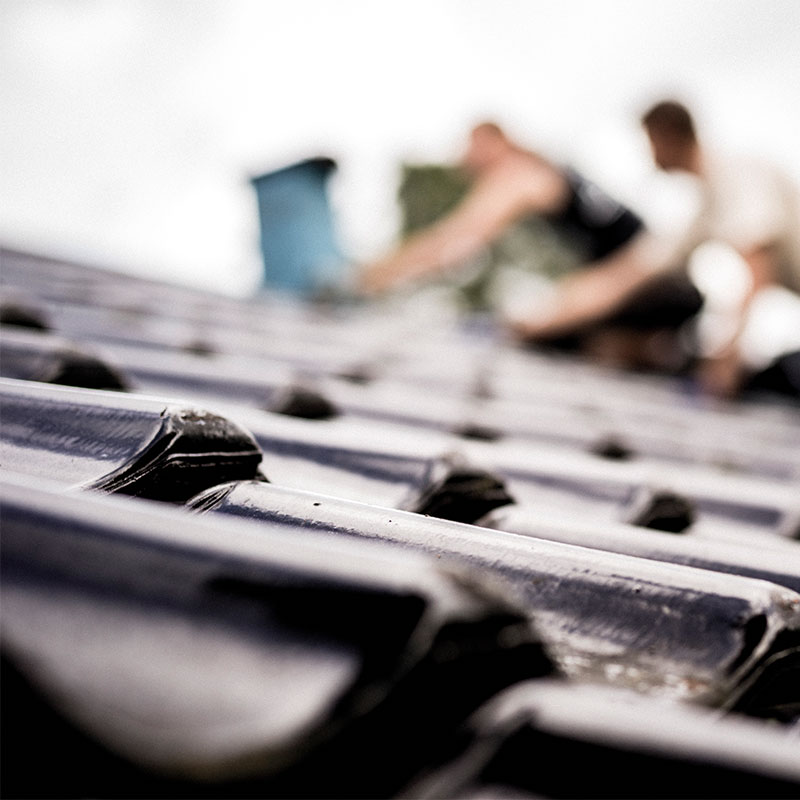Nærbilde av blanke, mørkeblå takstein mens to håndverkere legger tak i bakgrunnen.