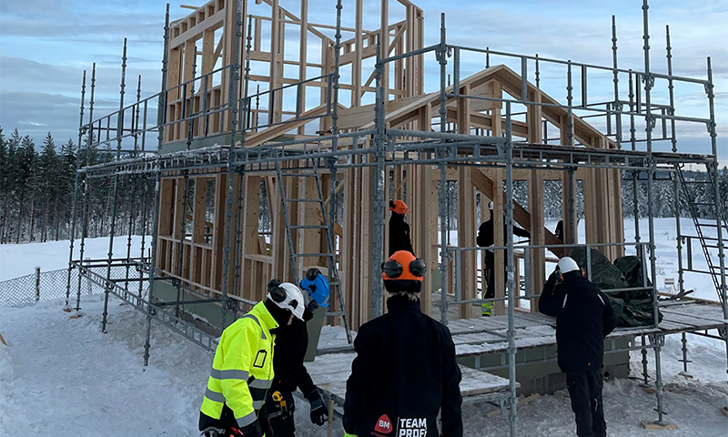 Bindingsverkhus i tre under bygging på snødekt tomt med grå stillas og arbeidere i fargerike hjelmer.