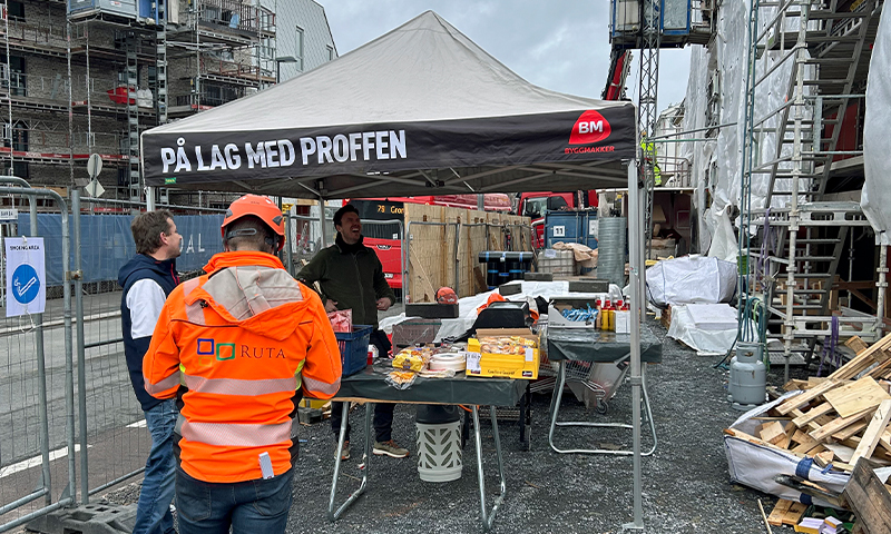Byggeplass med svart telt merket PÅ LAG MED PROFFEN, snacksbord og oransje jakker.