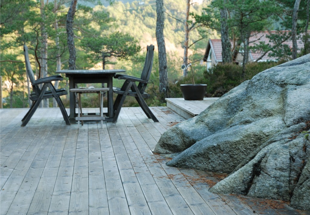 Uteplass i furuskog med grå terrasse, svart bord og stoler ved granittstein.