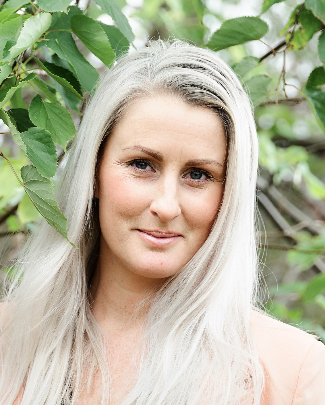 Person med langt sølvhvitt hår i ferskenfarget blazer foran grønn løvbakgrunn