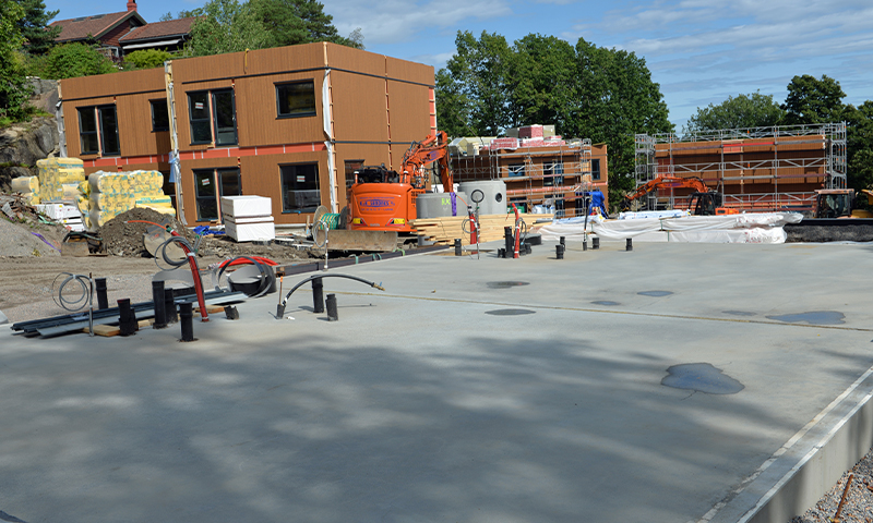 Byggeplass med grå betongplate i forgrunn, brune modulbygg, oransje gravemaskiner og rør