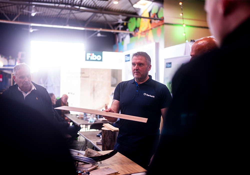 Mann i Tarkett-skjorte viser frem en gulvplanke på en messe med Fibo-stand i bakgrunnen.