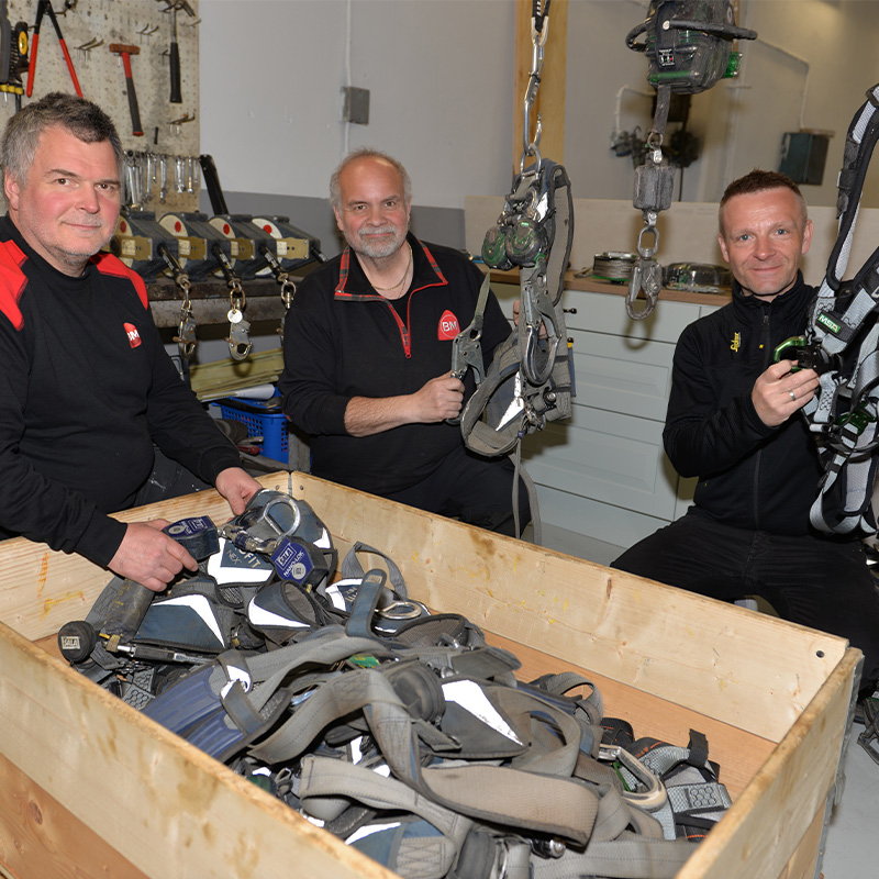 Tre menn i svarte arbeidstøy holder grå sikkerhetsseler over kasse med seler i verksted