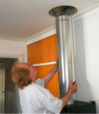 Mann i hvit t-skjorte monterer skinnende stålpipe mot hvit vegg og oransje skap.