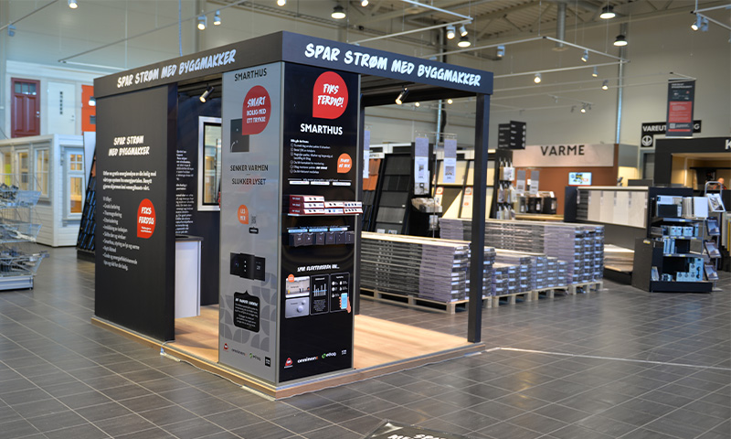 Svart- og rødfarget informasjonsstand med teksten “Spar strøm med Byggmakker” i lyst byggevarehus.
