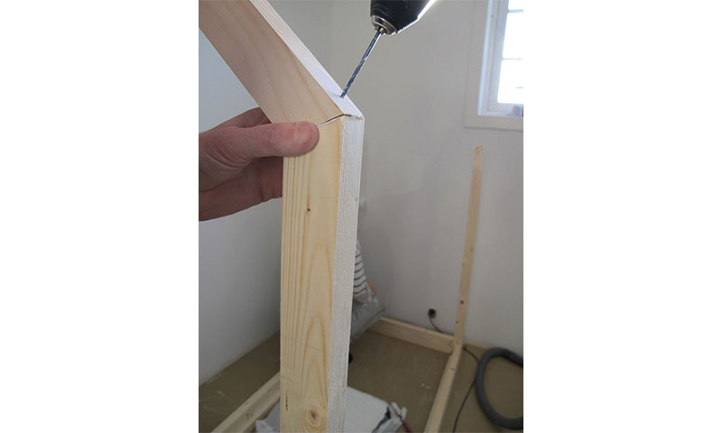 Hånd holder lyst furutre mens svart drill med blått bor borer i hjørnet av hvitt rom