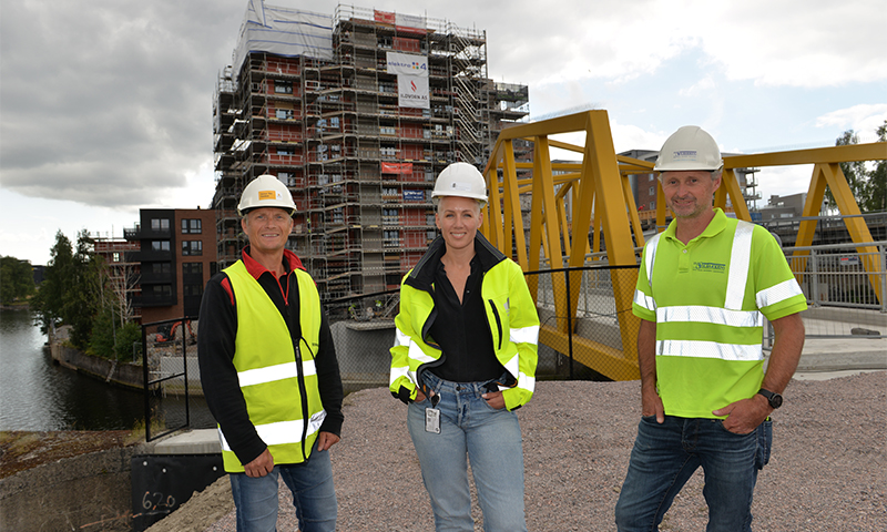 Tre byggearbeidere med grønne vernevester og hvite hjelmer foran stillasert boligbygg og gul bro