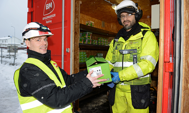 To anleggsarbeidere i gule jakker bytter grønn eske ved rød Byggmakker-kontainer i snølandskap.
