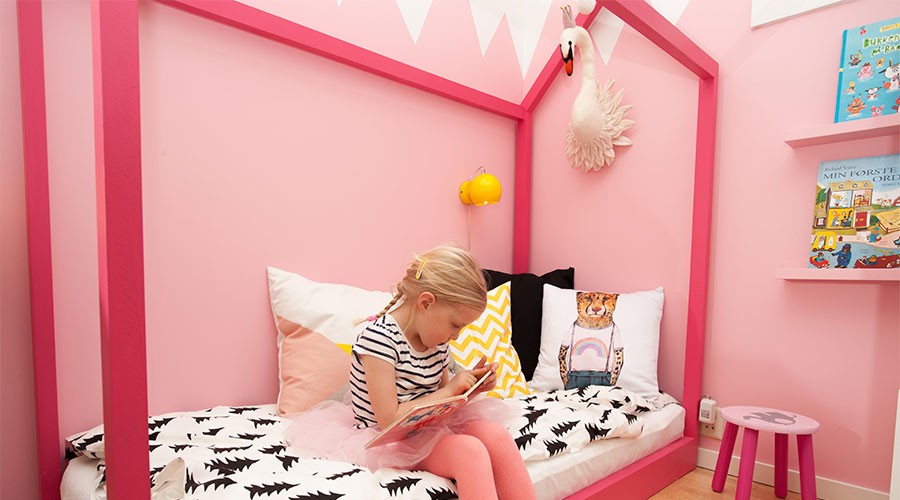 Jente leser bok på rosa barnerom med husformet seng, svart-hvitt sengetøy og fargerike puter