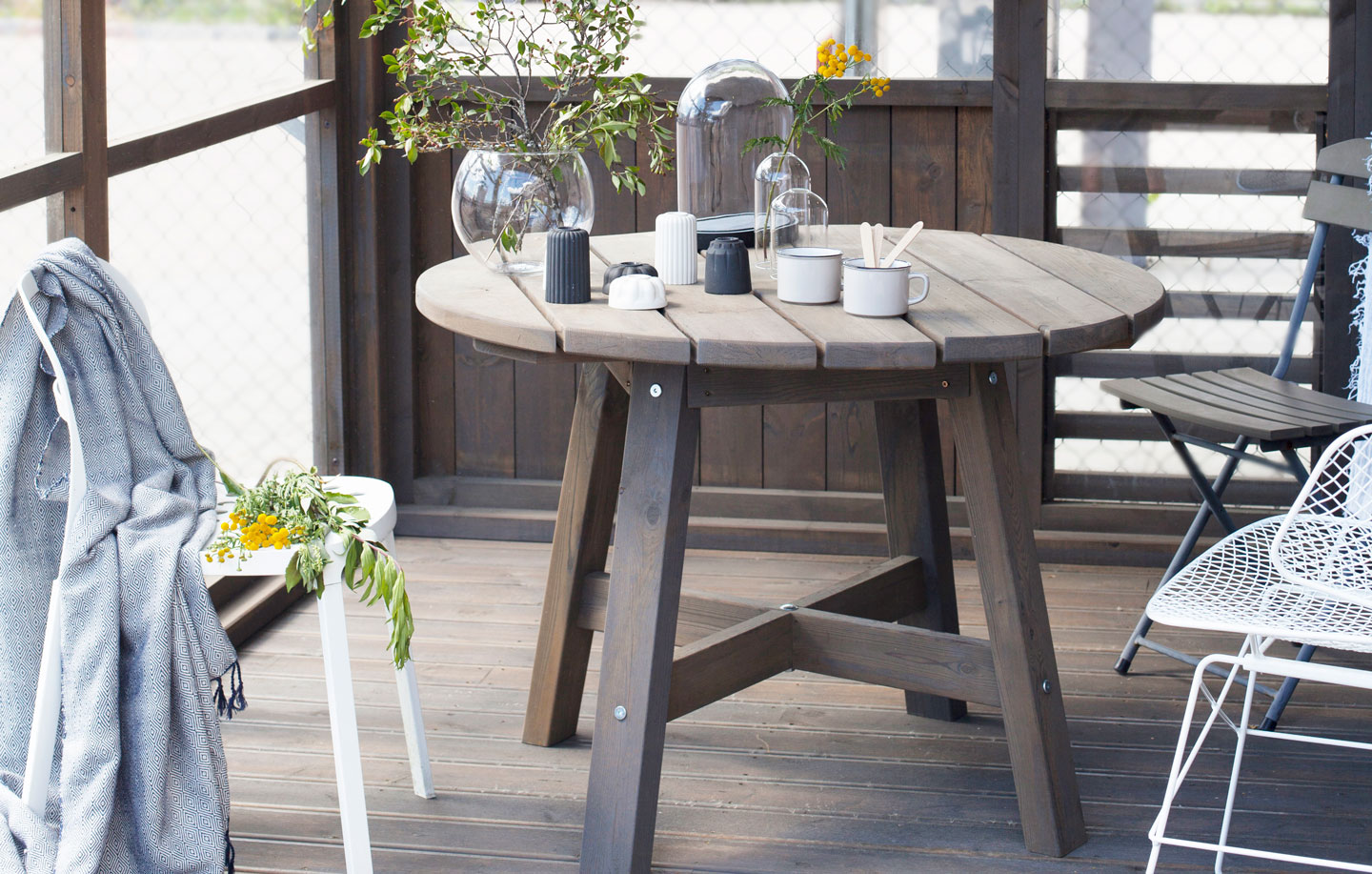 Rustikk veranda med rundt mørkt trebord, keramikkkrus, glassvaser, gul blomsterkvist og grå pledd.