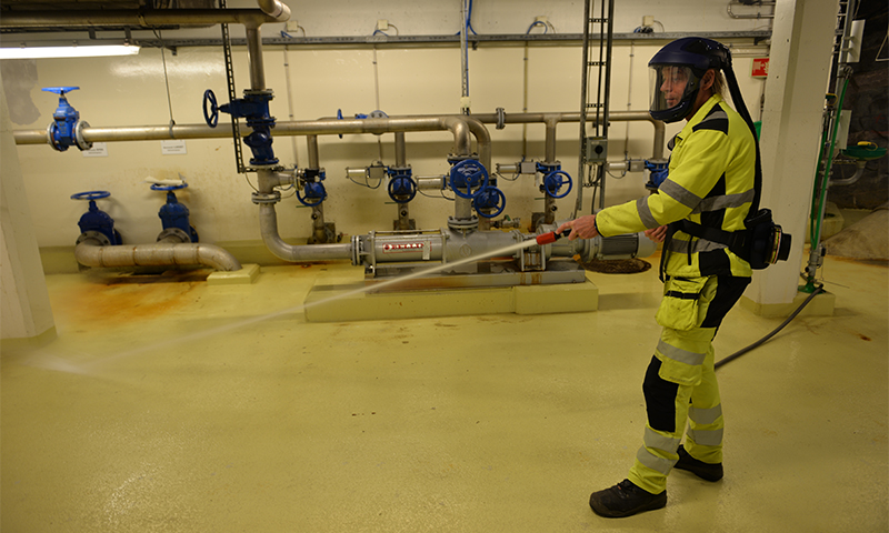 Person i gul varnedrakt spyler gulvet med høytrykksvann i maskinrom med blå ventiler.