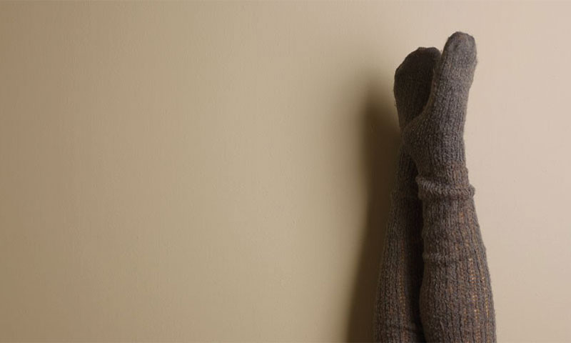 To kryssede ben i strikkede gråbrune sokker mot lys beige vegg.