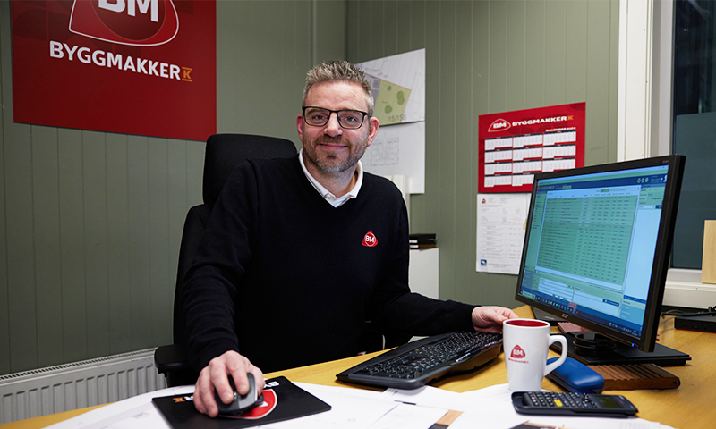 Kontorarbeider i mørk genser ved skrivebord med PC, kaffekopp og rød Byggmakker-logo på grønn vegg.