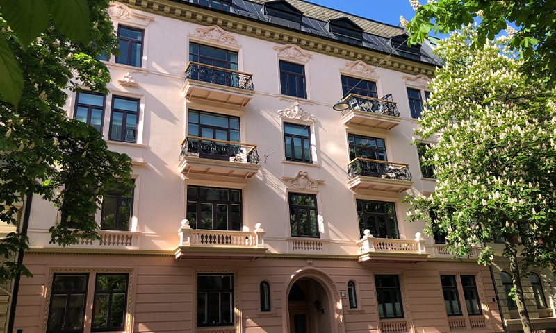 Pastellrosa bygård med smijernsbalkonger omgitt av grønne trær og blomstrende kastanjer.