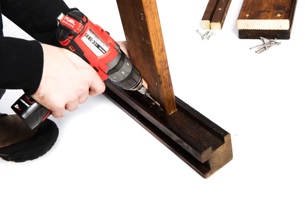 Hånd med rød-svart batteridrill skrur mørkebrunt trestykke på hvit bakgrunn.