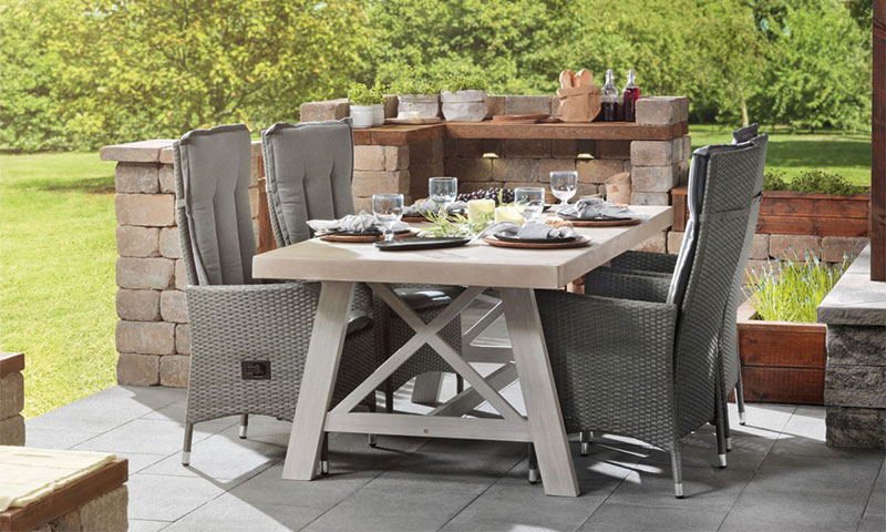 Utebord i hage: lyst trebord med fire grå kurvstoler, dekket med servise og vinglass.