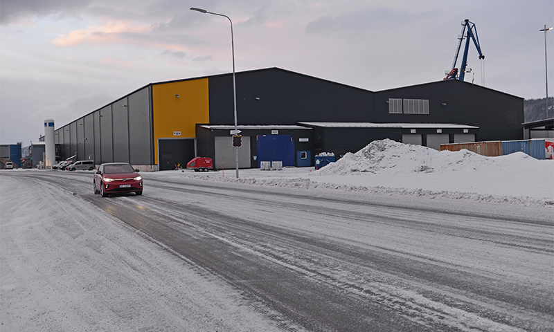 En rød bil kjører forbi svart-gult lagerbygg på snødekt vei med kran i bakgrunnen