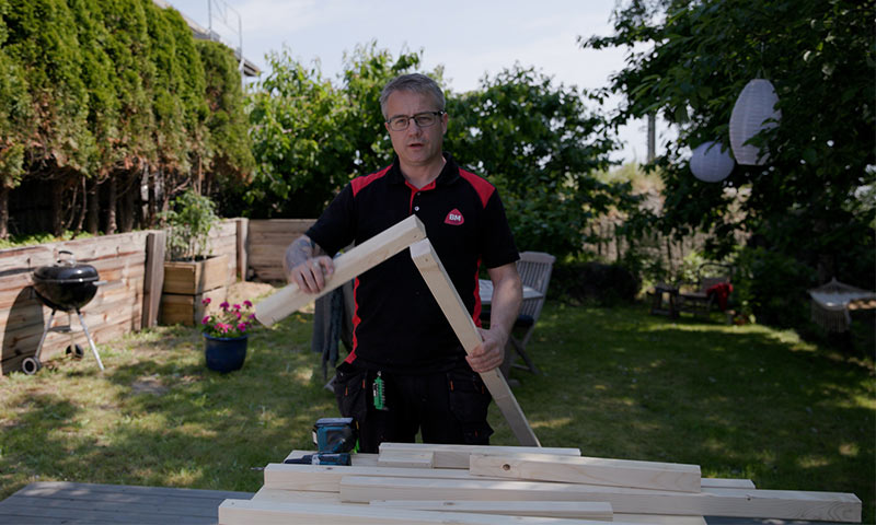 Håndverker i svart- og rød skjorte monterer treramme på arbeidsbenk i grønn hage.