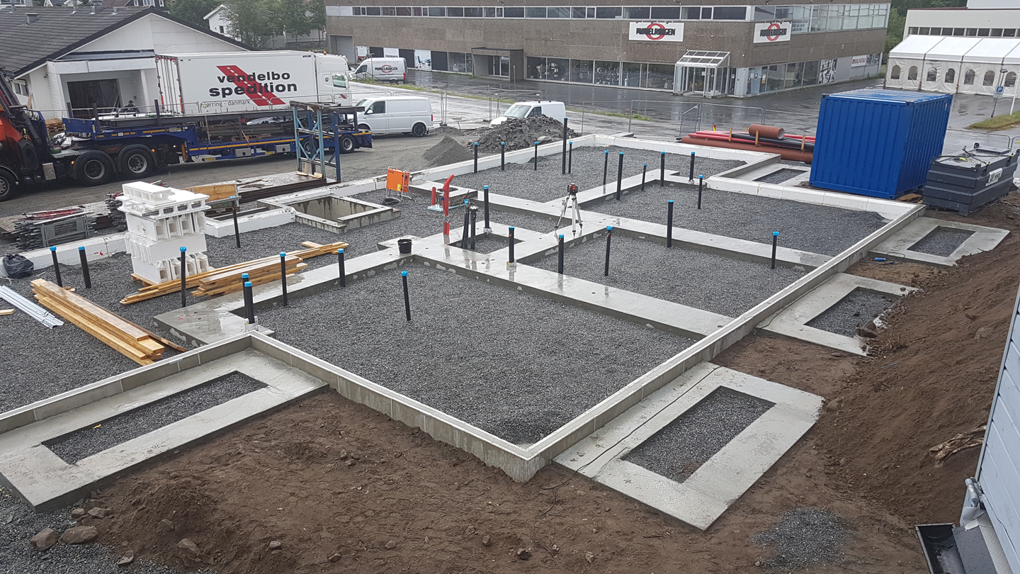 Regnvåt byggeplass med grå betongfundament, grus, blå rørstusser og blå container.