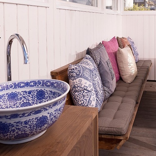 Utebenk med grå dyne og mønstrete puter i blått, rosa, beige ved blåhvit porselensvask