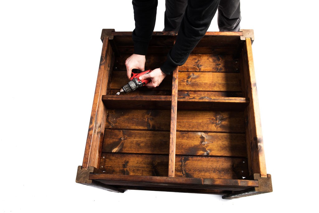 Person med svart genser skrur brun trekasse med rød drill på hvit bakgrunn.