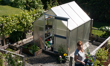 Lite glassdrivhus med metallramme i solfylt hage, omkranset av pallekarmer, kvinne vanner planter.