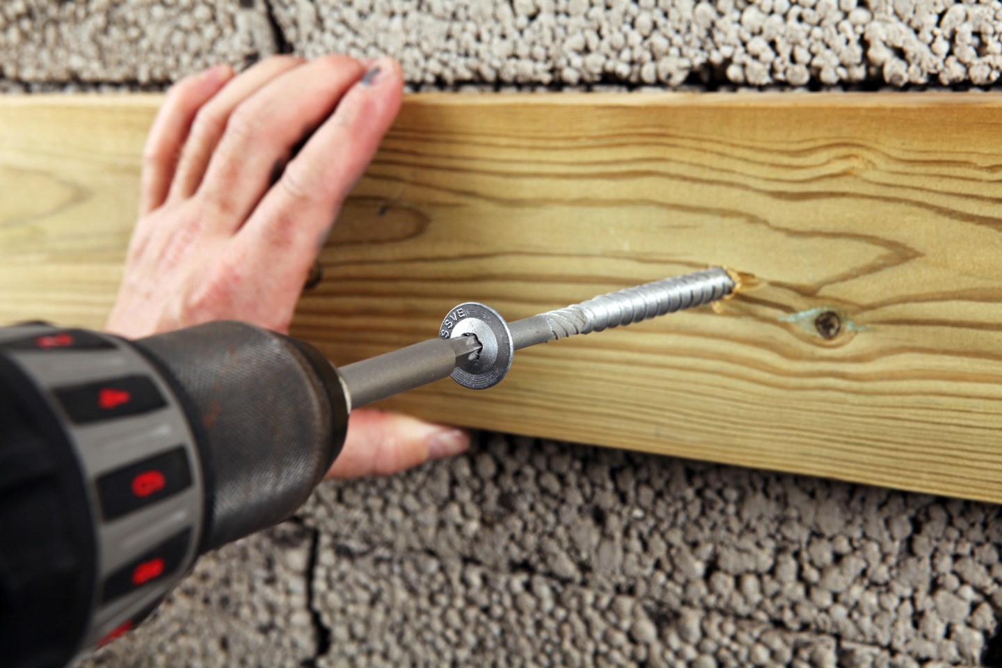 Nærbilde av drill som fester stålskrue i lyst treverk på grå betongvegg, hånd støtter
