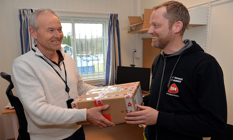 To personer i kontor skifter brun pappeske med rødt hjertetape ved skrivebord, blåstripete gardiner