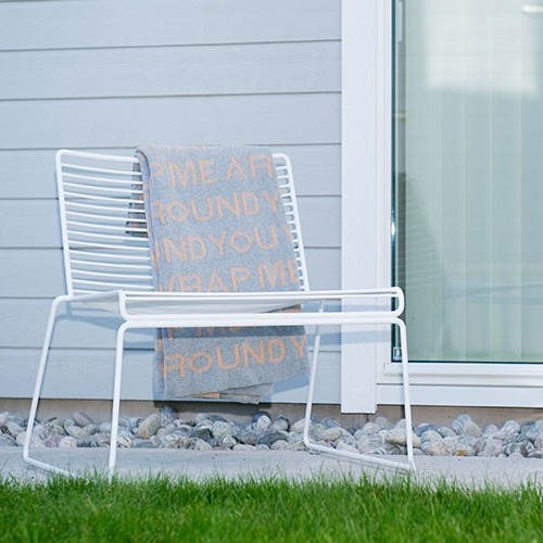 Hvit metallstol med grått pledd med oransje tekst foran grå husvegg og glassdør, stein og gress