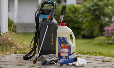 Svart ryggsprøyte og hvit beholder med blå-rød etikett ved malerull, brett og hansker på hageheller