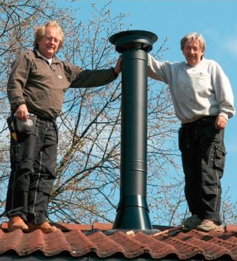 To håndverkere på rød takstein ved svart skorsteinpipe mot blå himmel.