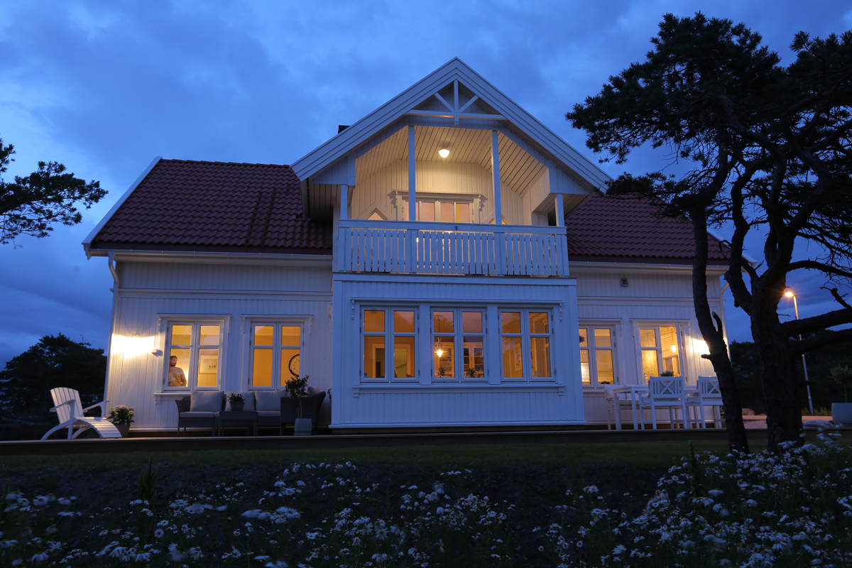 Hvit toetasjes trehus i skumring med varme lys, balkong, hagemøbler og blomster
