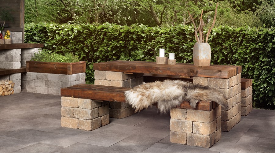 Utebord og benk av treplanker på steinblokker, skinn, grå steingulv og grønn hekk