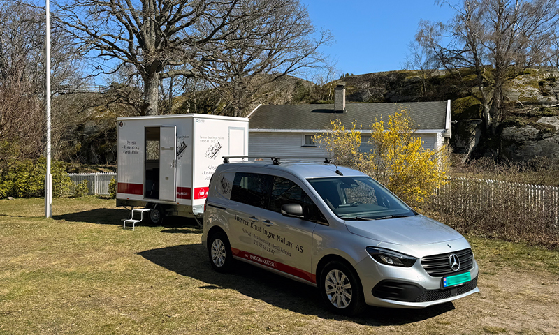 Sølv Mercedes-bil med Tømrer Nalum AS-logo foran hvit brakke, gule forsythiabusker