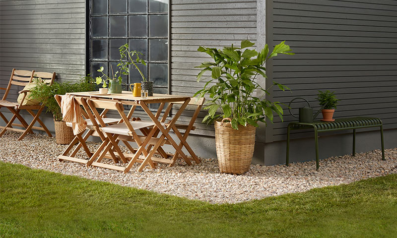 Uteplass ved lysegrå vegg med trebord, stoler og grønne planter i kurver på grus