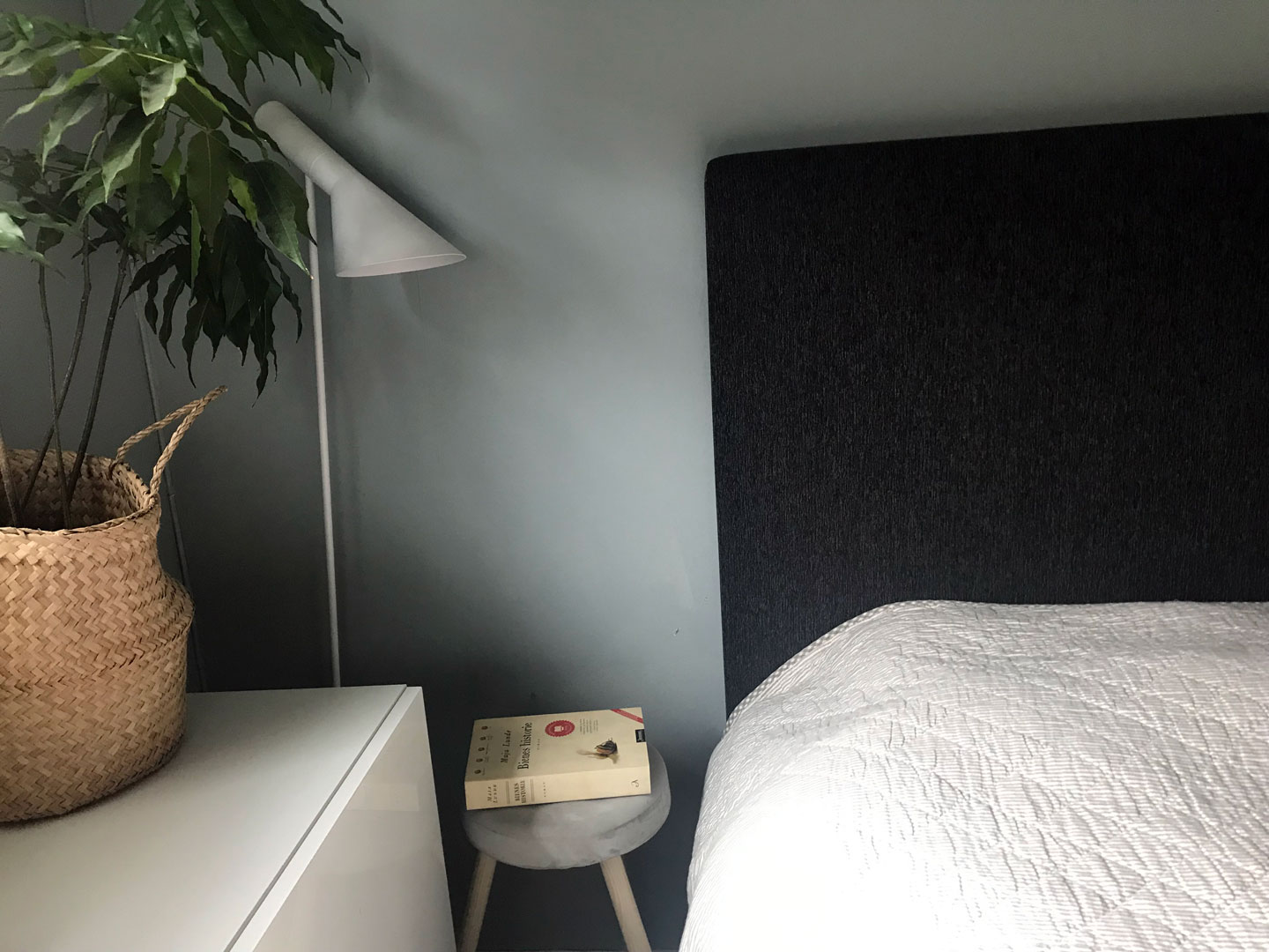 Soveromshjørne med mørk grå sengegavl, hvit dyne, rund pall med bok, kurv med grønt plante.