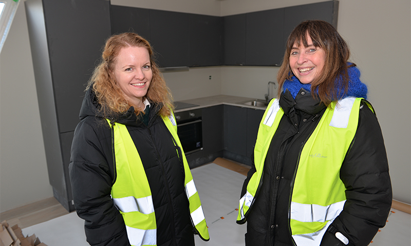 To kvinner i gule refleksvester foran moderne grått kjøkken med stekeovn og vask.