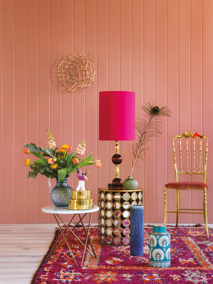 Interiør med rosa panelvegg, fargerikt persiskteppe, blomstervase, gullmøbler og rosa lampe.