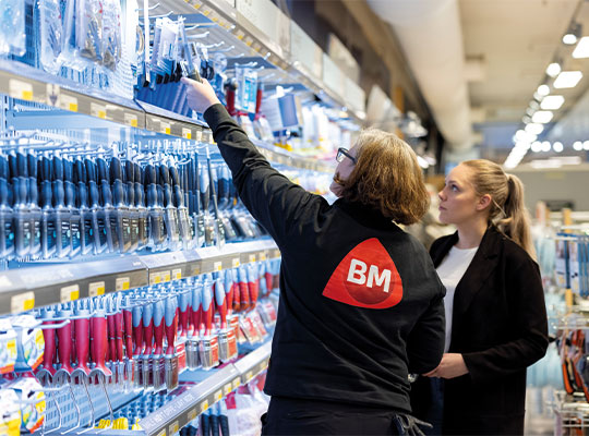 To ansatte i svart genser med rød BM-logo foran reol med blå og røde skrujern.