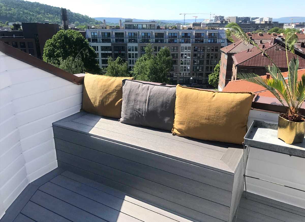 Takterrasse i sol med grå benk, sennepsgule og grå puter, palme og byutsikt
