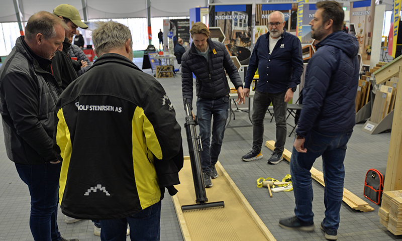 Fem menn på byggevaremesse, én demonstrerer svart håndverktøy på lys treflis på grått gulv.