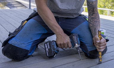 Nærbilde av arbeidshender i blå bukser som borer hull i grå terrasse med batteridrevet drill