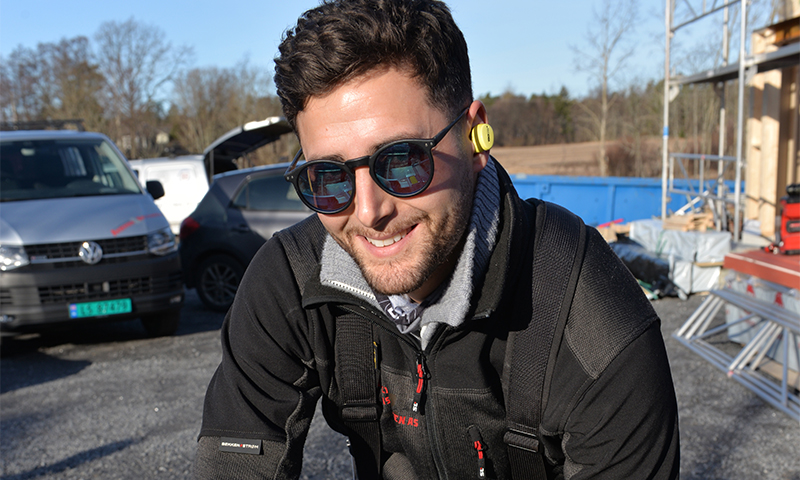 Person i svart jakke og gult hørselvern på byggeplass med biler, stillas og materialer.