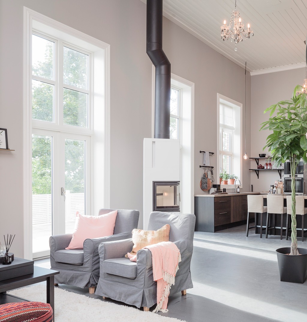 Lyst, åpent sturområde under høyt tak, to grå lenestoler med rosa puter foran hvit vedovn.