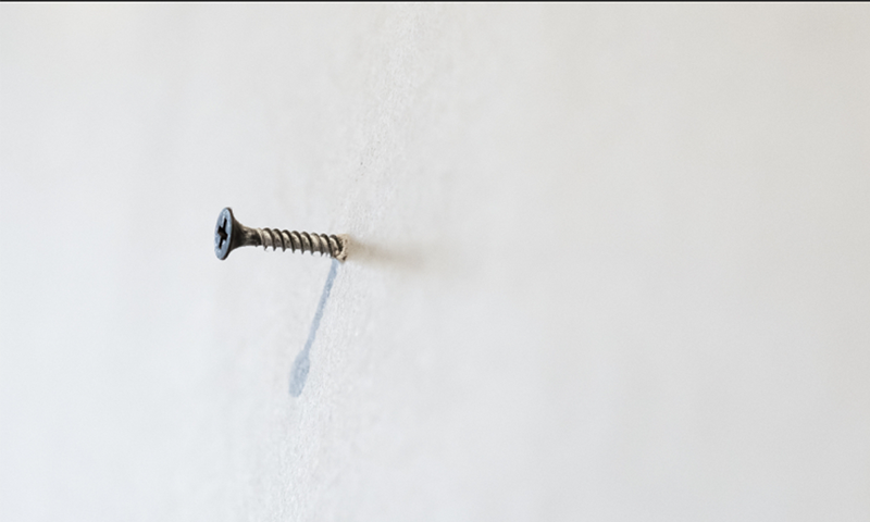 Nærbilde av metallskrue halvveis skrudd inn i ensfarget hvit vegg og skygge.